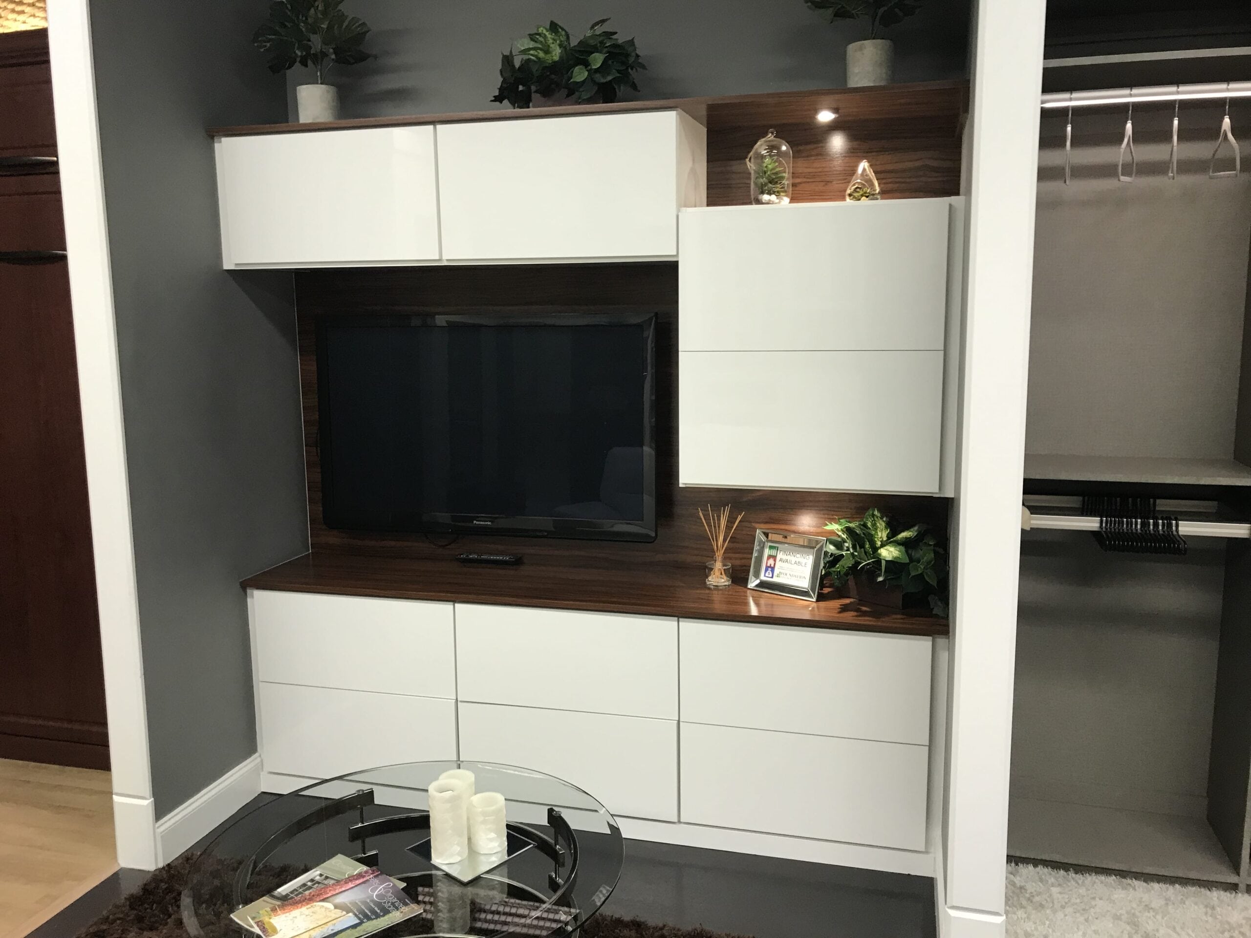 able-closet Modern living room setup with a flat-screen TV on a wooden cabinet, white storage units, plants, decorative items, and a glass coffee table with magazines and candles in front. Closet space is visible on the right.