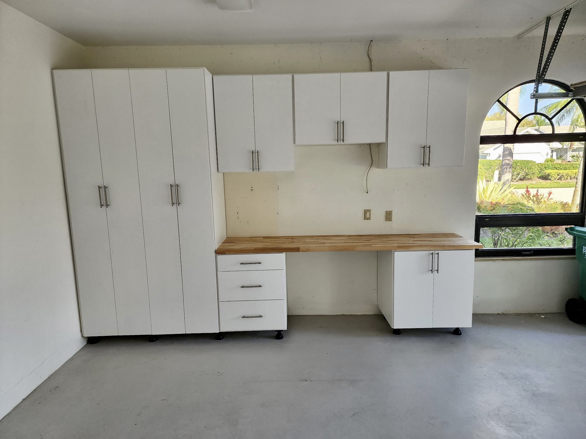 able-closet White cabinets with a wooden countertop and drawers are installed against a garage wall. Upper and lower storage units are present, with a window on the right showing a garden outside. The floor is smooth and gray.