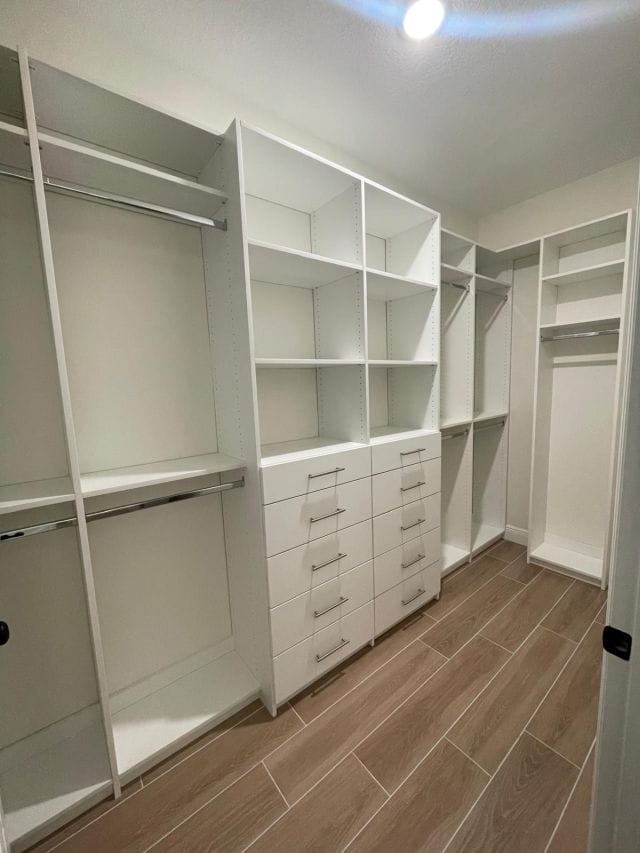 able-closet A spacious walk-in closet with white shelves, drawers, and hanging rods, featuring wood-look tile flooring and bright overhead lighting.