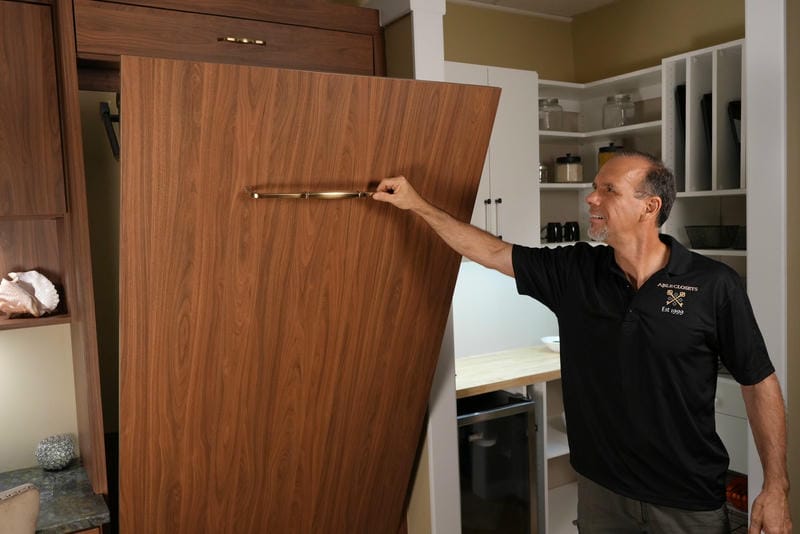 able-closet A man in a black shirt pulls down a vertical wooden Murphy bed in a modern room with shelves and cabinets in the background.