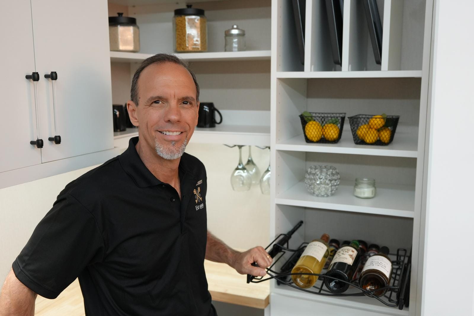 able-closet A man smiling and standing in a pantry, holding a sliding wine rack with bottles. Shelves behind him hold baskets of lemons, glasses, jars, and mugs. The man wears a black polo shirt.