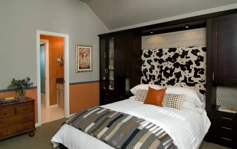 able-closet A modern bedroom with a bed featuring a black and white cow print headboard, white bedding, and striped throw. Dark wood murphy beds and cabinets surround the bed. A dresser with flowers and books sits to the left, near a doorway to the bathroom.