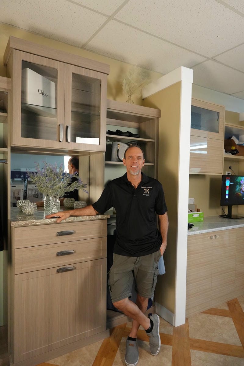 able-closet A man in a black shirt and shorts leans against a kitchen counter, smiling. The kitchen has light wood cabinets, a TV in the background, and decorative items on the counter.