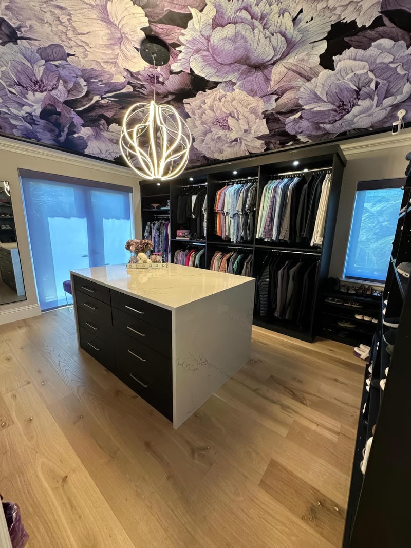 able-closet A modern walk-in closet with black shelving, a central marble-topped island, organized clothes, large windows, wood floors, and a bold purple floral ceiling design. Stylish pendant lighting hangs from the center.