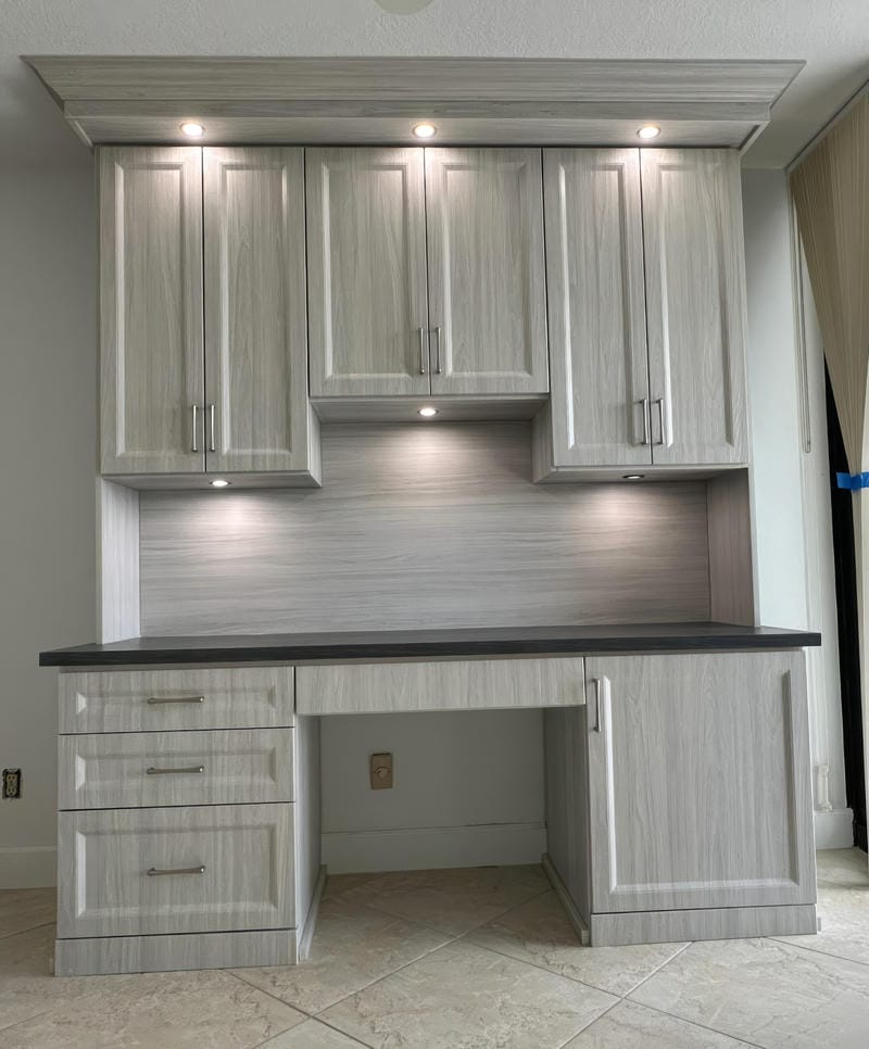 able-closet Modern built-in desk with light gray wood cabinets, drawers, and under-cabinet lighting. The desk has a black countertop, tiled floor, and is set against a light-colored wall. Curtains partially cover the window on the right.