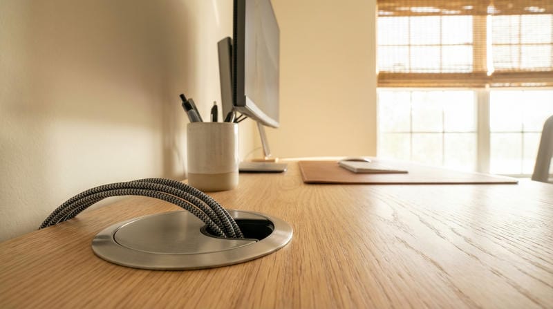 able-closet A close-up of a tidy wooden desk with a cable grommet, computer monitor, keyboard, mouse, and a cup holding pens, set in a bright room with large windows and bamboo blinds.