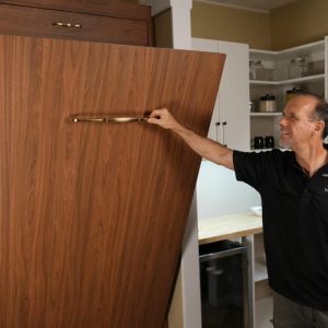 able-closet A man in a black shirt pulls down a vertical wooden Murphy bed in a modern room with shelves and cabinets in the background.