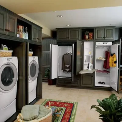 able-closet A spacious laundry room features two front-loading washers and dryers, abundant laundry room storage with built-in cabinets, a closet with ironing board, hanging clothes, a desk with computer, houseplants, and a colorful rug on tiled flooring.