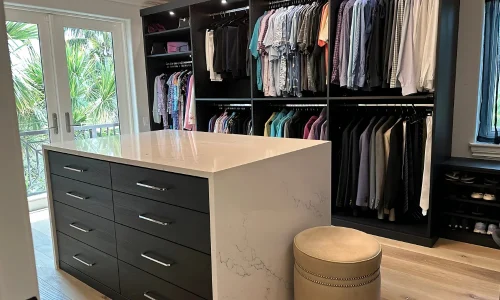 able-closet A modern walk-in closet features organized hanging clothes, a large white island with drawers, a round beige ottoman, and natural light streaming in from glass doors showing greenery outside.