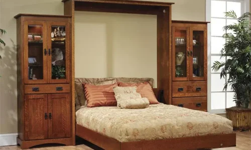 able-closet A wooden murphy bed with a light brown bedspread is flanked by two matching cabinets with glass doors and shelves, set against a beige wall with a plant near a window.