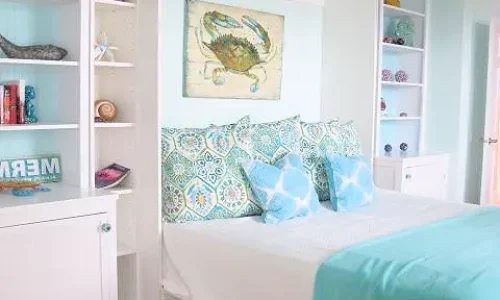 able-closet A bright, coastal-themed bedroom features open white shelves, a murphy bed with patterned blue and green pillows, turquoise bedding, and crab artwork above. Decor includes seashells, books, and other beachy accents.