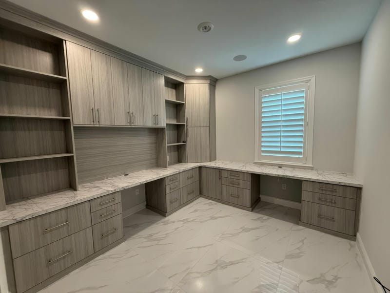 able-closet Modern home office with built-in wood cabinets, open shelves, and marble-patterned countertops, featuring tiled floors and a window with white plantation shutters. Room is well-lit with recessed ceiling lights.