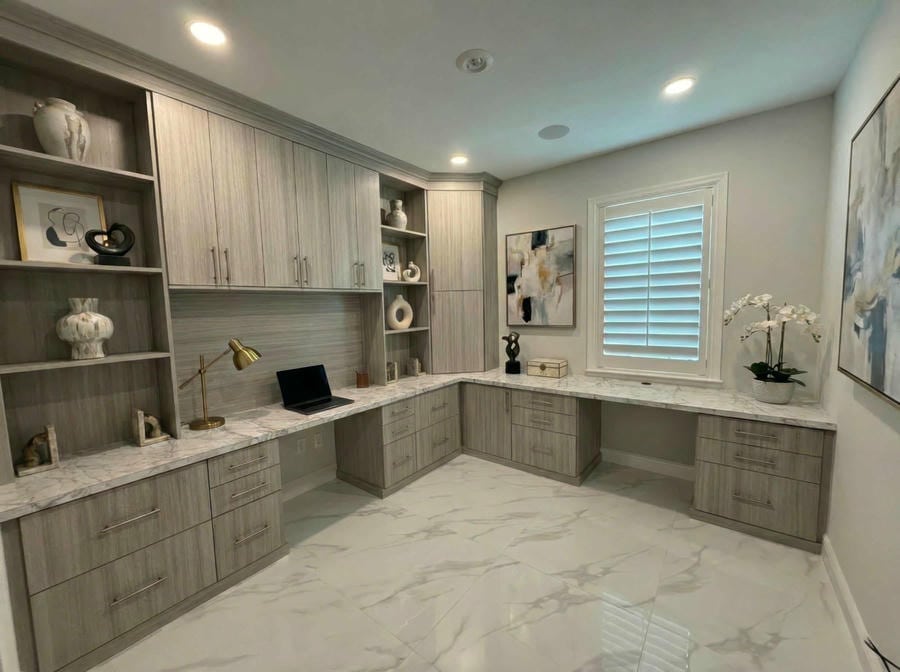 able-closet Modern home office with marble tile floor, L-shaped marble desk, built-in light wood cabinets, shelves with decorative objects, a laptop, brass desk lamp, window with white shutters, and abstract wall art.