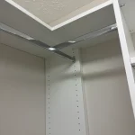 able-closet A close-up view of a white closet corner with two chrome hanging rods meeting at a right angle, mounted to the walls and shelves under a textured ceiling.
