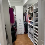 able-closet A neatly organized walk-in closet with wooden floors, white shelves, drawers, hanging clothes, handbags, shoes, and storage baskets, well-lit by a ceiling light.