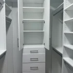 able-closet A white built-in closet with open upper cabinets, five shelves, and four closed drawers below. The closet has additional shelves and hanging rods on either side, and the floor is wooden.
