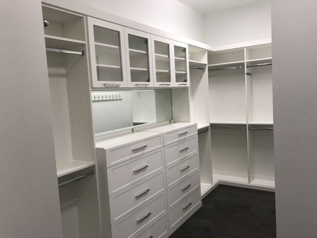 able-closet A spacious walk-in closet with white shelving, drawers, glass-front cabinets, and multiple hanging rods, all set against dark wood flooring and white walls.
